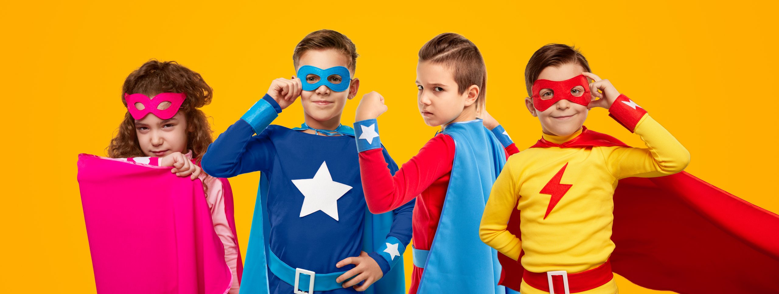 Company of brave kids in colorful superhero capes and masks lookin at camera on yellow background in studio